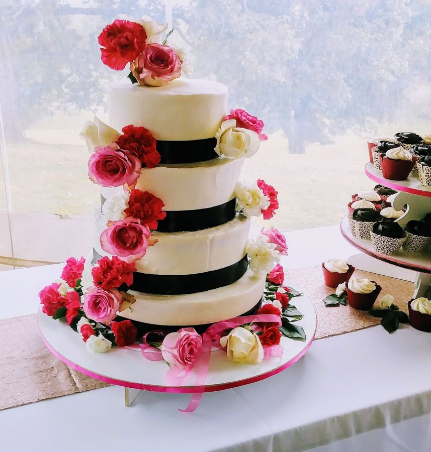 Four-tier white wedding cake with black ribbon and cascading pink and red roses, with cupcakes